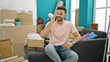 Genç İspanyol adam akıllı telefondan konuşuyor. Yeni evindeki kanepede oturuyor.