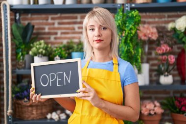 Young caucasian woman working at florist holding open sign relaxed with serious expression on face. simple and natural looking at the camera. 