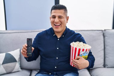 İspanyol genç adam patlamış mısır yiyor televizyon kumandası kullanarak kameraya seksi bir ifadeyle bakıyor, neşeli ve mutlu bir yüzle.. 