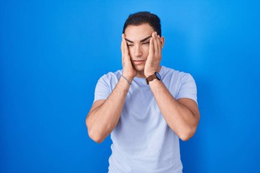Young hispanic man standing over blue background tired hands covering face, depression and sadness, upset and irritated for problem 