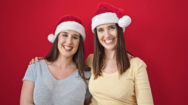 Birbirlerine sarılan iki kadın izole edilmiş kırmızı arka planda noel şapkası takıyorlar.