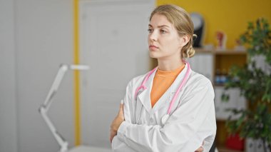 Genç sarışın kadın doktor, klinikte ciddi bir ifade ve kollarını kavuşturarak duruyor.