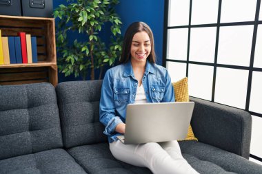 Evde dizüstü bilgisayar kullanan İspanyol kadın pozitif ve mutlu görünüyor. Dişlerini gösteren kendine güvenen bir gülümsemeyle ayakta duruyor ve gülümsüyor. 