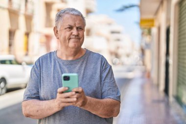 Orta yaşlı, kır saçlı bir adam sokakta rahat bir ifadeyle akıllı telefon kullanıyor.