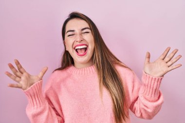 Young hispanic woman standing over pink background celebrating mad and crazy for success with arms raised and closed eyes screaming excited. winner concept 