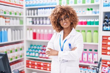 Afro-Amerikalı kadın eczacı gülümseyerek eczanede kollarını kavuşturup duruyor.