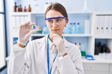 Bilim adamlarının laboratuvarında çalışan beyaz bir kadın bitcoin taşıyor ciddi bir yüz ifadesiyle eli çenesinde soruyu düşünüyor, kafa karıştırıcı bir fikir. 
