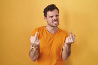Young hispanic man standing over yellow background showing middle finger doing fuck you bad expression, provocation and rude attitude. screaming excited 