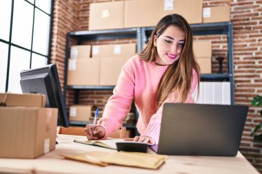 Young beautiful hispanic woman ecommerce business worker using laptop and calculator writing on notebook at office