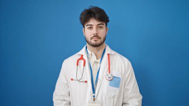 İzole edilmiş mavi arka planda ciddi bir ifadeyle duran genç İspanyol doktor.