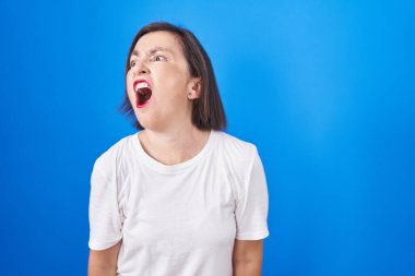 Middle age hispanic woman standing over blue background angry and mad screaming frustrated and furious, shouting with anger. rage and aggressive concept. 