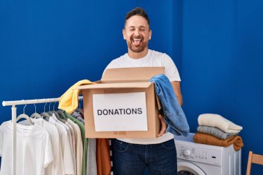 Orta yaşlı beyaz bir adam bağış kutusu tutuyor. Dilini çıkartıyor. Komik bir ifadeyle.. 