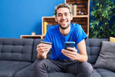 Evdeki kanepede oturan akıllı telefon ve kredi kartı kullanan genç adam.