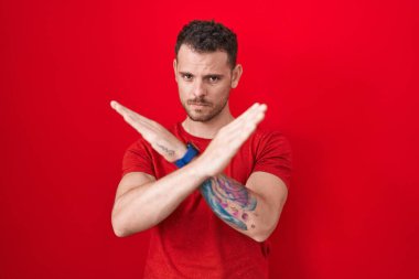 Young hispanic man standing over red background rejection expression crossing arms doing negative sign, angry face 