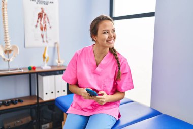 Genç ve güzel İspanyol fizyoterapist. Rehabilitasyon kliniğinde akıllı telefon kullanarak gülümsüyor.