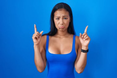 Hispanic woman standing over blue background pointing up looking sad and upset, indicating direction with fingers, unhappy and depressed. 