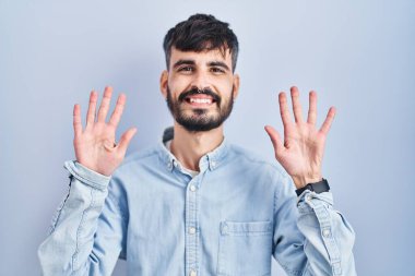 Mavi arka planda duran sakallı genç İspanyol adam on numarayı gösterip işaret ederken kendinden emin ve mutlu bir şekilde gülümsüyor.. 