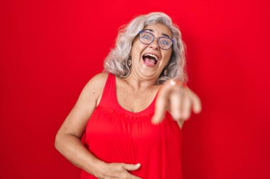 Kırmızı arka planda gri saçlı orta yaşlı bir kadın sana gülüyor, eli vücudunun üzerinde kamerayı işaret ediyor, utanç ifadesi var. 