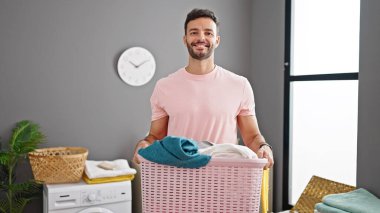 Çamaşır odasında çamaşır sepetini tutan kendine güvenen İspanyol genç adam.