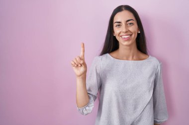 Genç esmer kadın pembe arka planda duruyor. Bir numarayı işaret ediyor ve gülümsüyor. Kendinden emin ve mutlu.. 