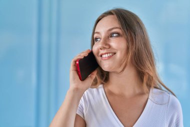 Sarışın, kendine güvenen genç bir kadın izole edilmiş mavi duvar arka planında akıllı telefondan konuşuyor.