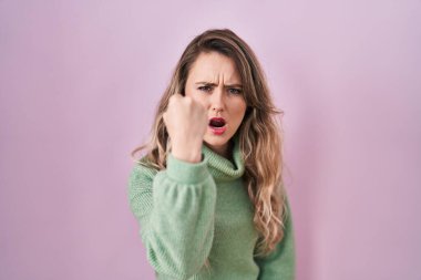 Young caucasian woman standing over pink background angry and mad raising fist frustrated and furious while shouting with anger. rage and aggressive concept. 