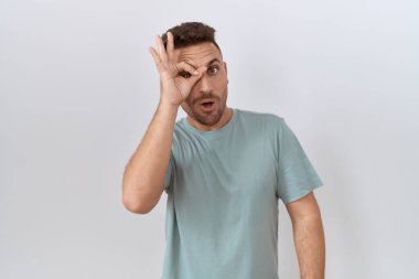 Hispanic man with beard standing over white background doing ok gesture shocked with surprised face, eye looking through fingers. unbelieving expression. 