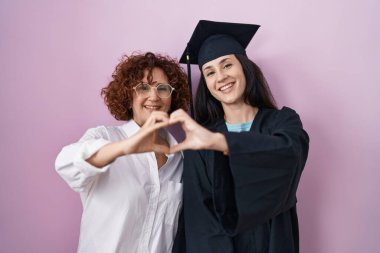 İspanyol anne ve kızı mezuniyet şapkası ve tören cübbesi takıyor. Sevgiyle gülümsüyor. Elleriyle kalp sembolü yapıyorlar. romantik kavram. 
