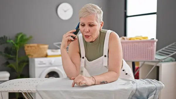 Middle age grey-haired woman talking on smartphone leaning on ironing board at laundry room