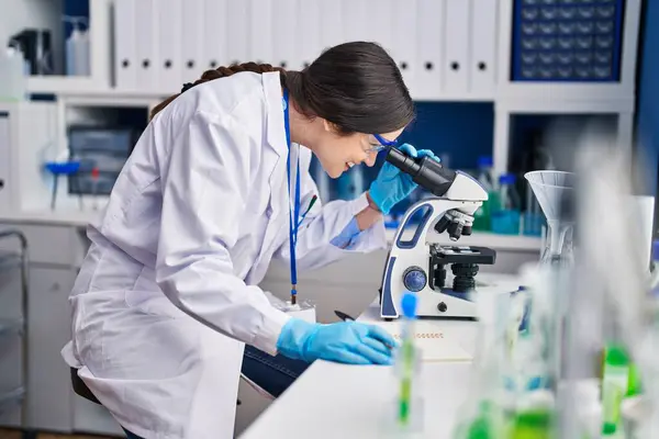 Young woman scientist writing on notebook using microscope at laboratory