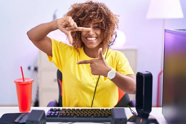 Young hispanic woman with curly hair playing video games wearing headphones smiling making frame with hands and fingers with happy face. creativity and photography concept.