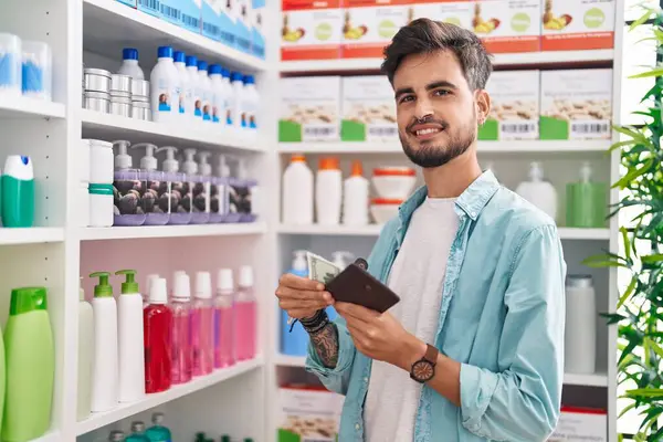 Young hispanic man customer smiling confident holding dollars at pharmacy