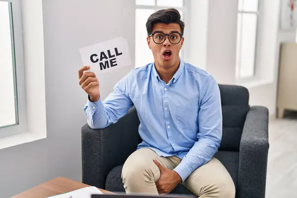 Hispanic man working at therapy office holding call me banner scared and amazed with open mouth for surprise, disbelief face