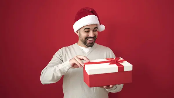 クリスマスの帽子を身に着けている若いヒスパニック男性は 孤立した赤い背景上の贈り物を解放します — ストック写真