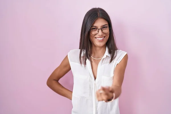 Brunette young woman standing over pink background wearing glasses beckoning come here gesture with hand inviting welcoming happy and smiling