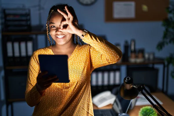 有辫子的非洲裔美国女人晚上在办公室里用平板电脑工作 手拿着手势 面带微笑 用手指看着她 面带微笑 — 图库照片