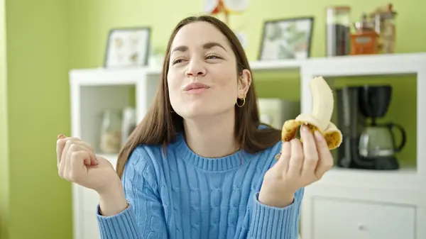 Young caucasian woman eating banana sitting on table at dinning room