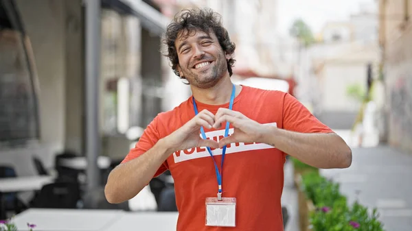 Young hispanic man activist wearing volunteer uniform doing heart gesture at street