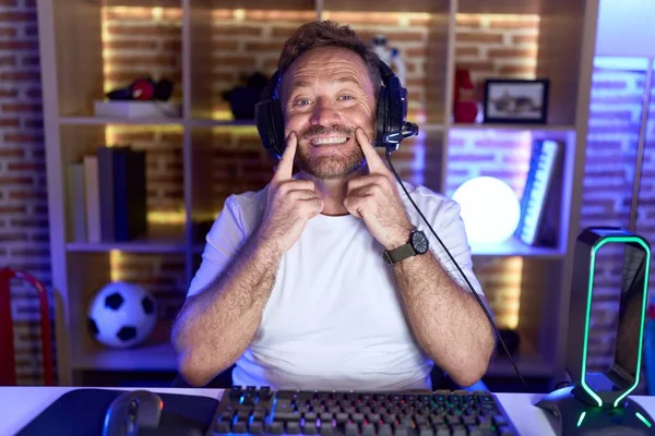 Middle age man with beard playing video games wearing headphones smiling with open mouth, fingers pointing and forcing cheerful smile