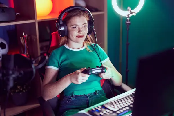 Young redhead woman streamer playing video game using joystick at gaming room