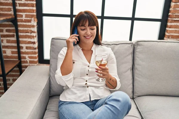 Young beautiful hispanic woman talking on the smartphone and drinking wine at home