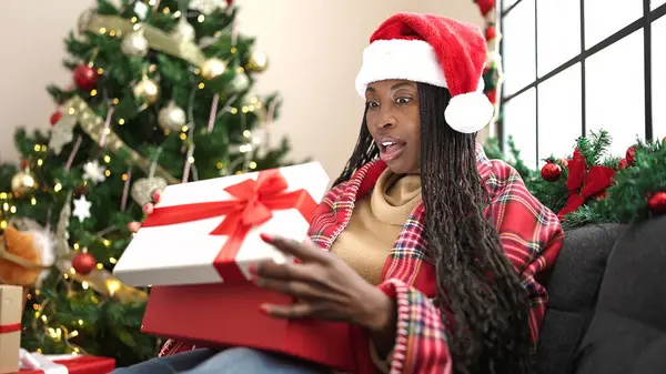 African woman unpacking gift sitting by christmas tree at home