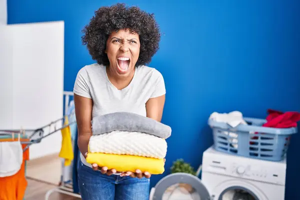 Black woman with curly hair holding clean laundry angry and mad screaming frustrated and furious, shouting with anger looking up.