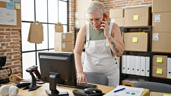 中年の灰色の髪の女性Eコマースビジネスワーカーは オフィスで働くコンピュータを使用してスマートフォンで話します — ストック写真