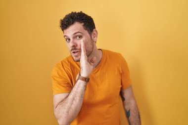 Young hispanic man standing over yellow background hand on mouth telling secret rumor, whispering malicious talk conversation 
