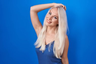 Mavi arka planda duran beyaz kadın. Kendine güvenen, el hareketleriyle saçlara dokunan, çekici ve modaya uygun poz veren. 