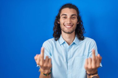 Young hispanic man standing over blue background showing middle finger doing fuck you bad expression, provocation and rude attitude. screaming excited 