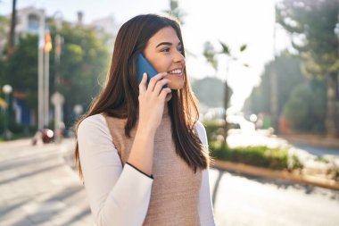Genç, güzel, İspanyol bir kadın gülümsüyor ve caddede akıllı telefondan konuşuyor.