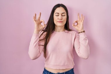 Genç esmer kadın pembe arka planda duruyor rahatlamış ve gözleri kapalı gülümsüyor parmaklarıyla meditasyon hareketi yapıyor. yoga konsepti. 