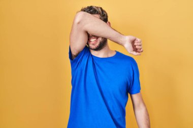 Sarı arka planda dikilen sakallı İspanyol bir adam. Gözleri kollarıyla kapatıyor. Neşeli ve komik gülümsüyor. kör kavram. 